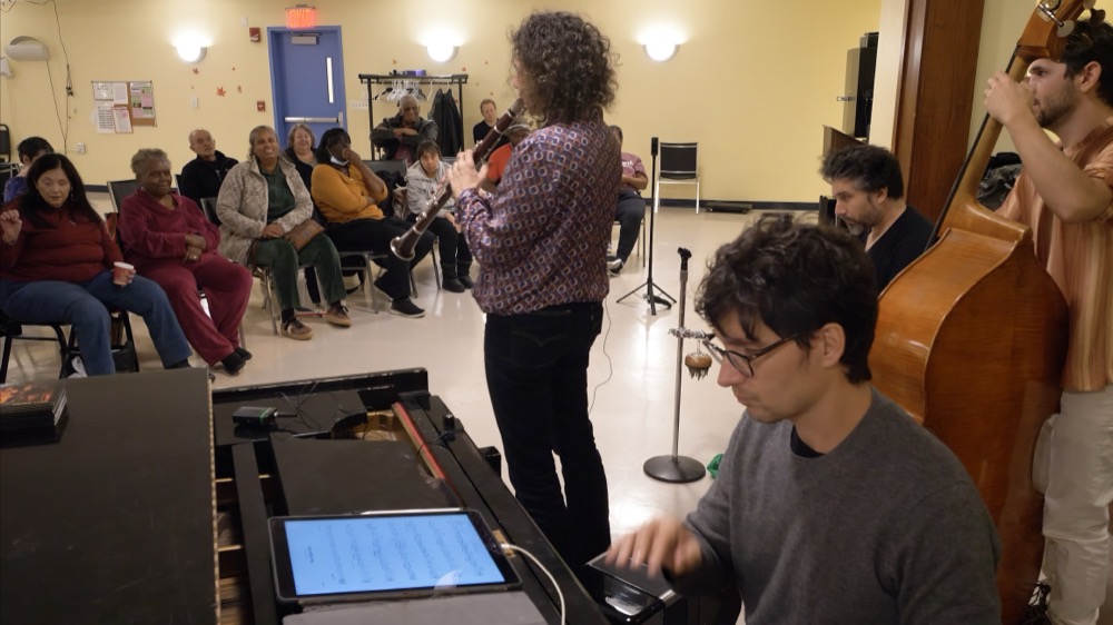 Anat Cohen plays clarinet for smiling listeners seated in a semicircle in a community room, a pianist and upright bassist behind her.