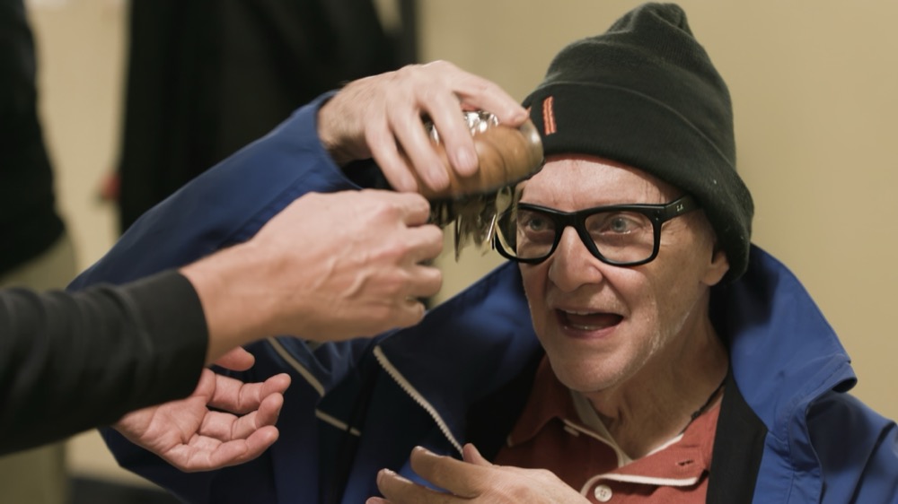An older man in a beanie and glasses beams as a musician holds a jingling shaker up to his ear so he can hear it up close.